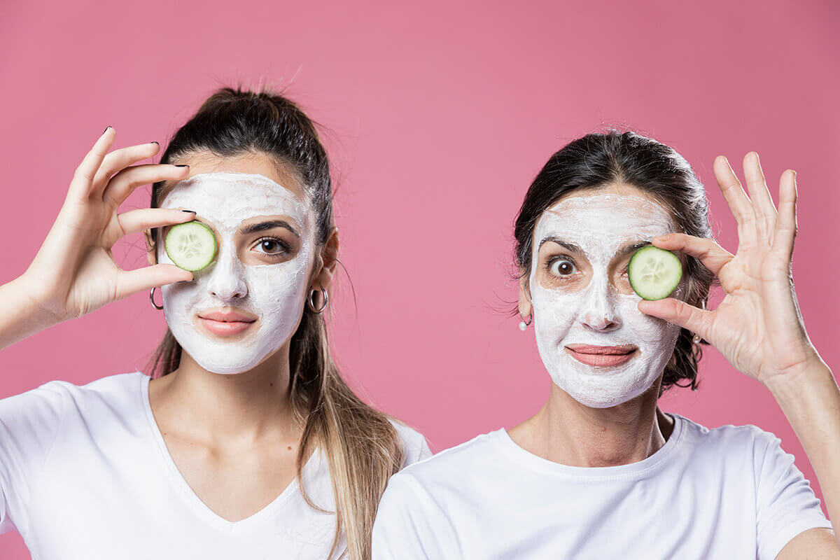 Две радостные женщины разного возраста на одном фото с нанесенными на кожу белыми (скорее всего из белой глины каолин) масками и колечком огурца