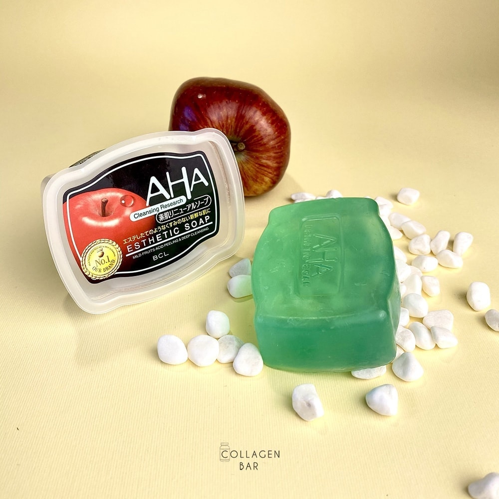 Упаковка і шматок мила BCL AHA Esthetic Soap на білих камінцях і червоне яблуко на задньому плані