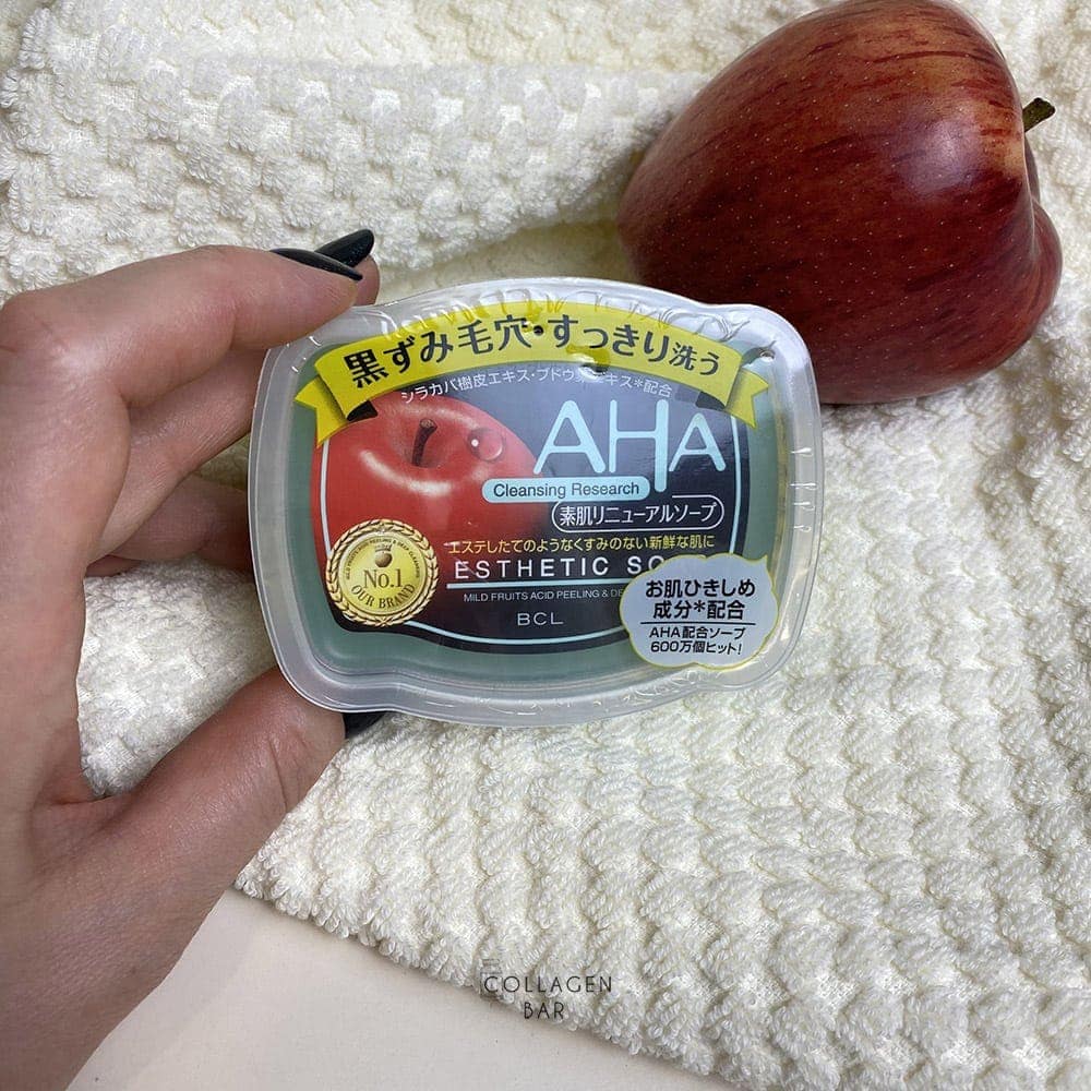 Упаковка мила BCL AHA Esthetic Soap у жіночій руці на тлі білого рушника і червоного яблука на рушнику