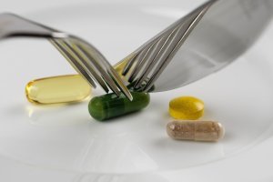 Різнокольорові таблетки та капсули на білій тарілці, над якими занесені ніж та вилка