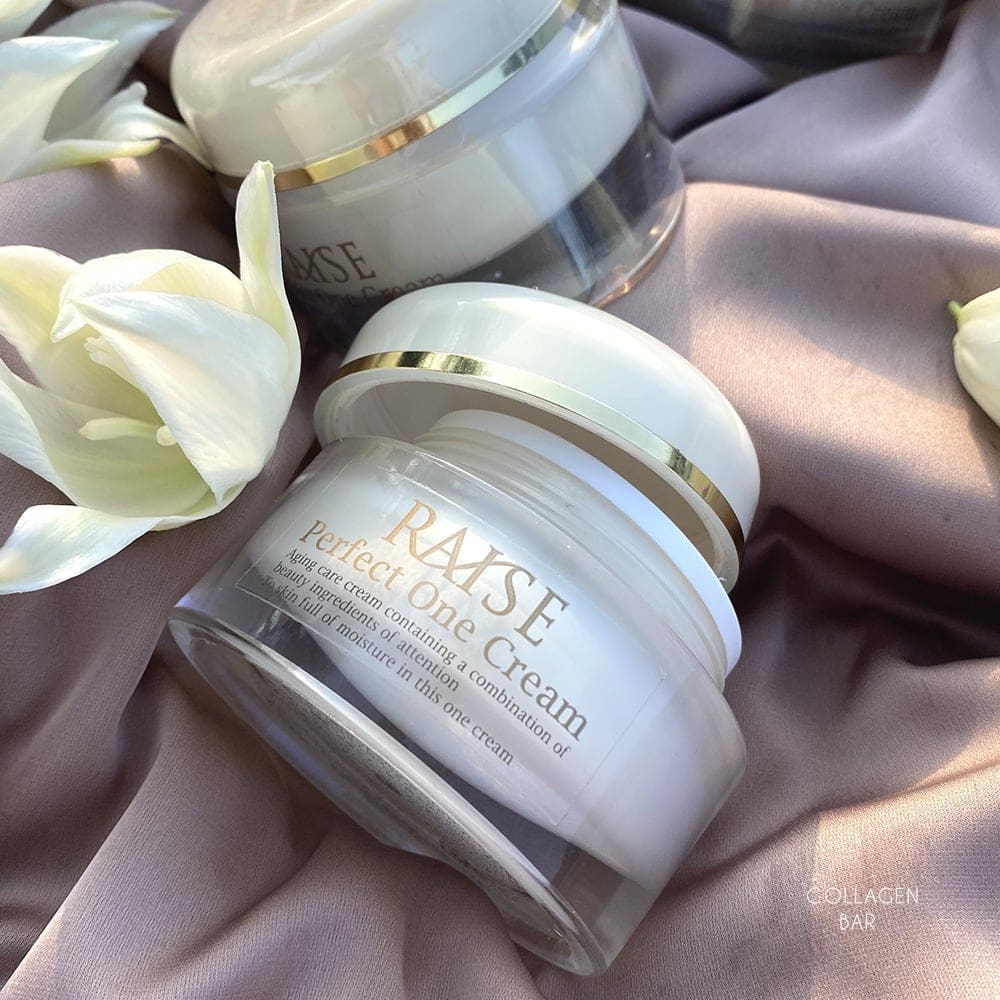 Дві баночки крему RAISE Perfect One Cream на світло-коричневій тканині з білою квіткою