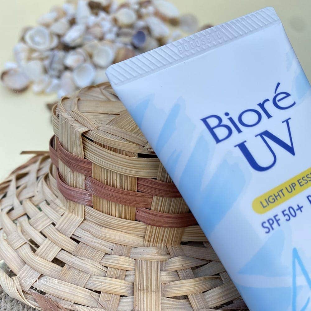 Фрагмент тюбика з санскрином Biore UV Light UP Essence із солом'яним капелюхом і морськими черепашками на задньому плані