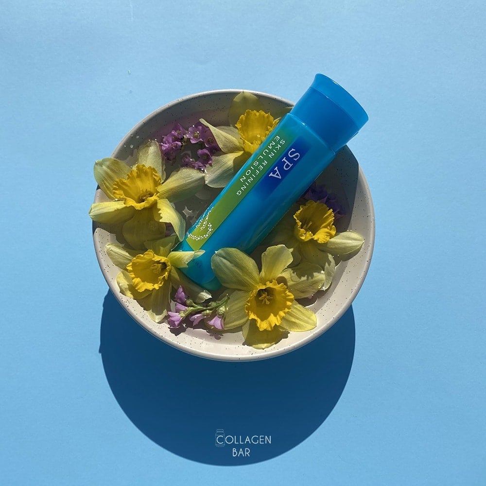 Эмульсия Hanajirushi SPA Skin Refining Emulsion в круглой пиале с цветками нарцисса и бадана на голубом фоне