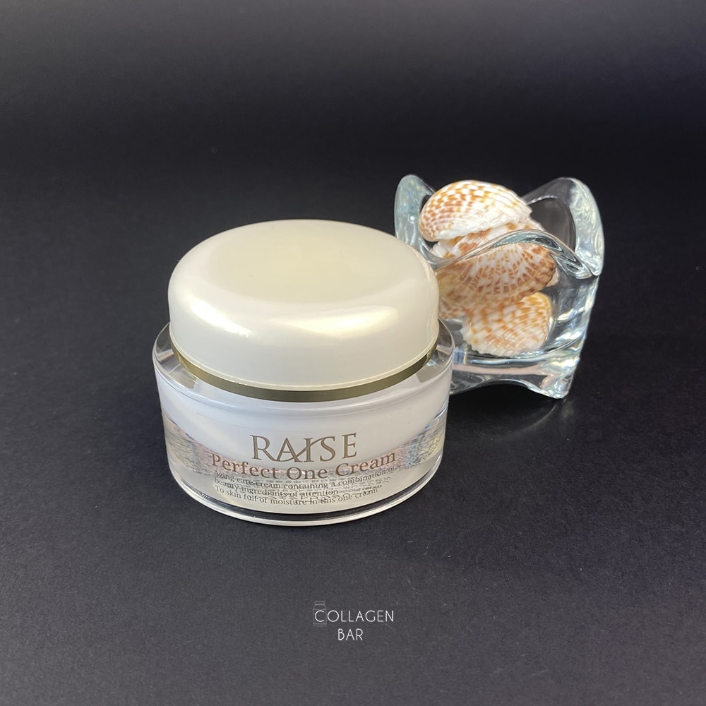 Антивіковий крем для обличчя RAISE Perfect One Cream на чорному тлі зі скляним прозорим свічником із морськими мушлями
