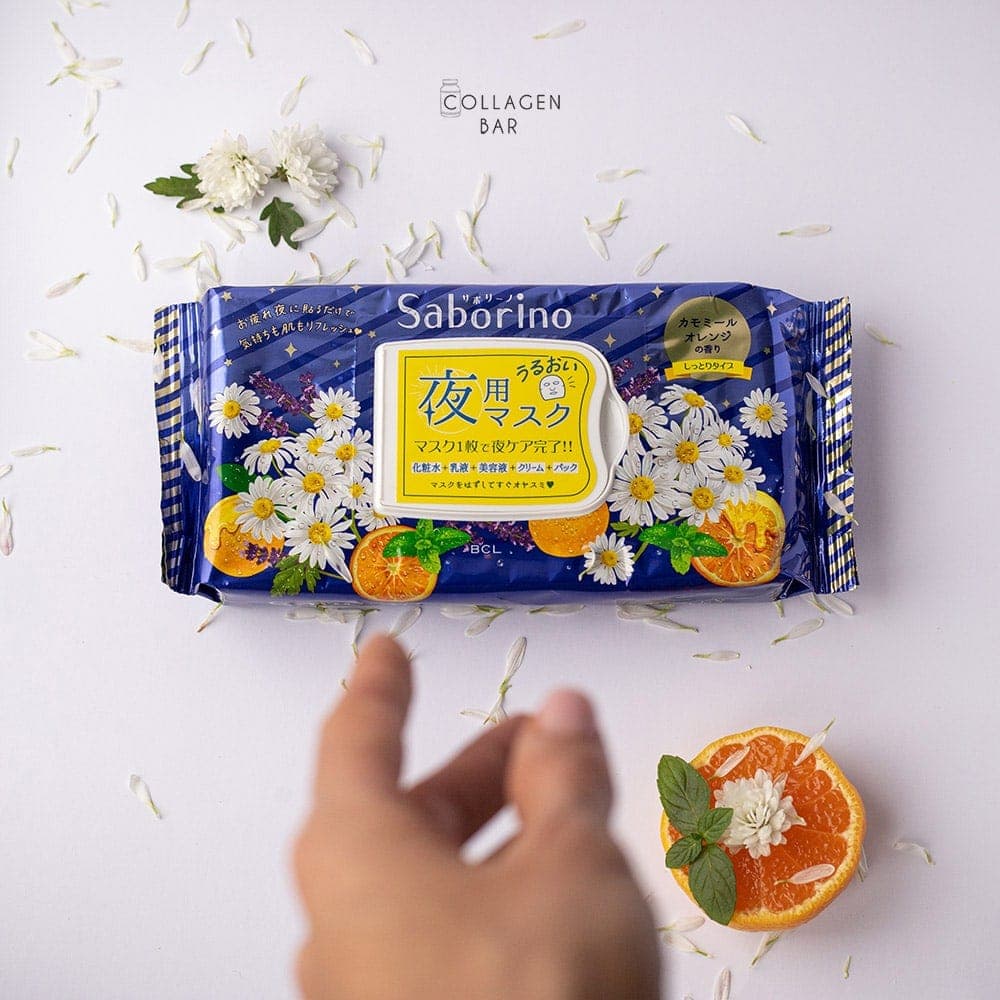 Вечірня маска Saborino tiredness mask з половиною апельсина, білими квітковими пелюстками і зеленими листочками м'яти та простягнута до неї жіноча рука