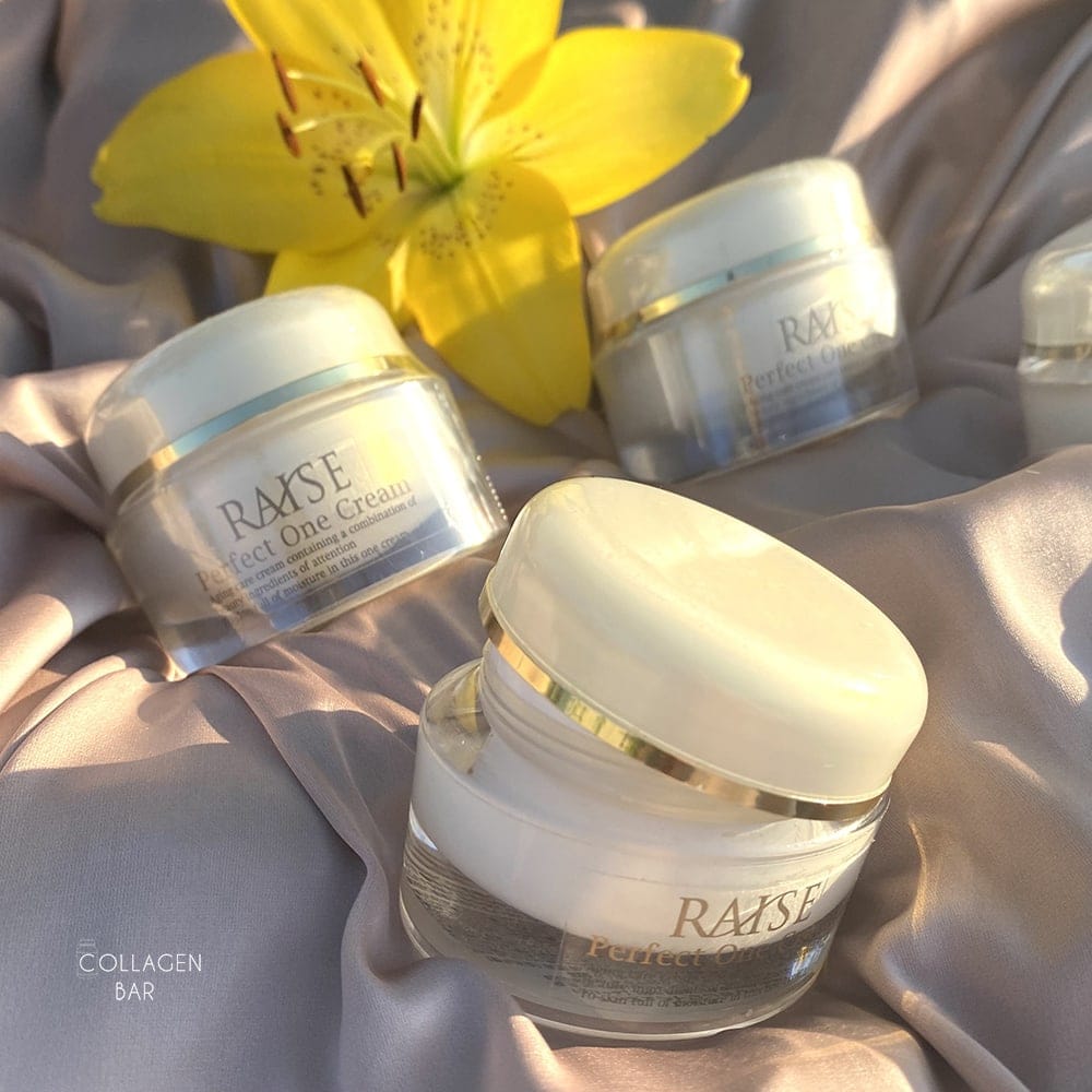 Три баночки крему RAISE Perfect One Cream на світло-коричневій тканині з квіткою жовтого лілійника