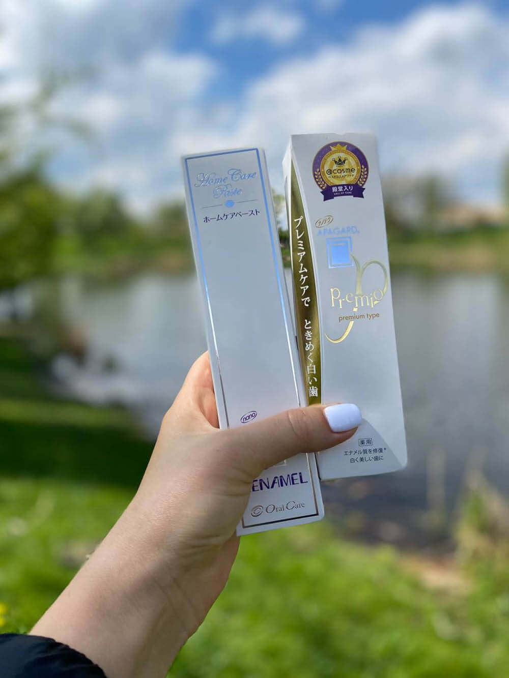 Зубные пасты с гидроксиапатитом Apagard Premio и Apagard Renamel в женской руке с белым маникюром на фоне пейзажа в расфокусе