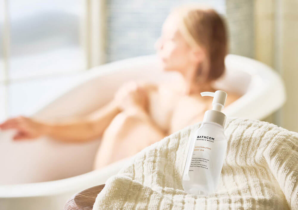 Дівчина приймає ванну на задньому плані в розфокусі й олія BATHCOM Moisturizing Body Oil на рушнику на передньому плані. Фото взято з сайту виробника за адресою: https://www.bathcom.jp/